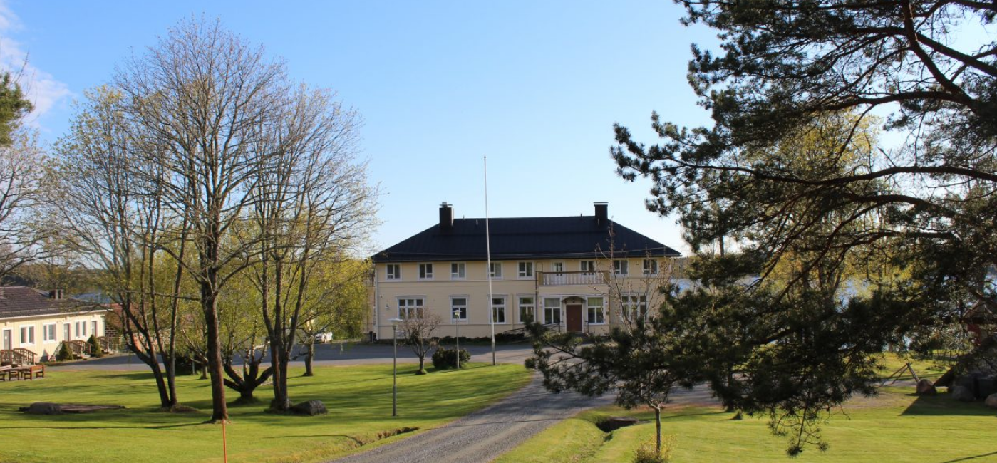 Ett stort gult trähus vid gräsplan och i bakgrunden strand.