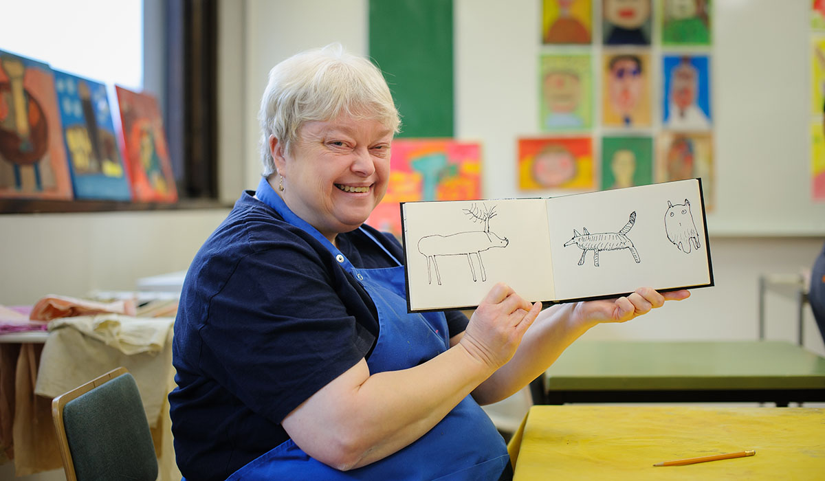 En äldre kvinna som visar upp sina teckningar på djur. I bakgrunden syns färggranna målningar och andra konstverk.
