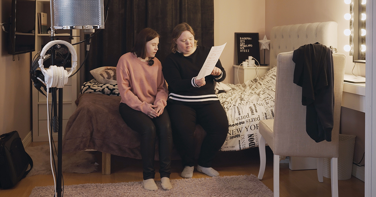 Två unga kvinnor med Downs syndrom sitter på säng med papper i handen.
