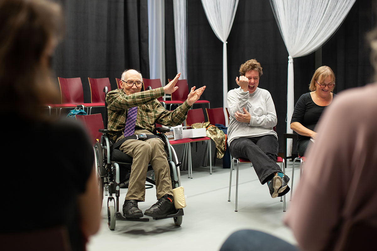 Skrattande äldre man i rullstol med händerna uppe och bredvis honom en kvinna som skrattar hjärtligt.