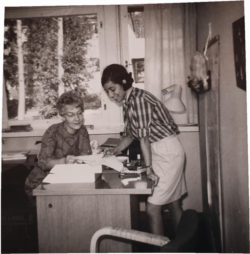 Svartvitt foto på två kvinnor i 60-talslook kollar tillsammans på dokument vid ett skrivbord.