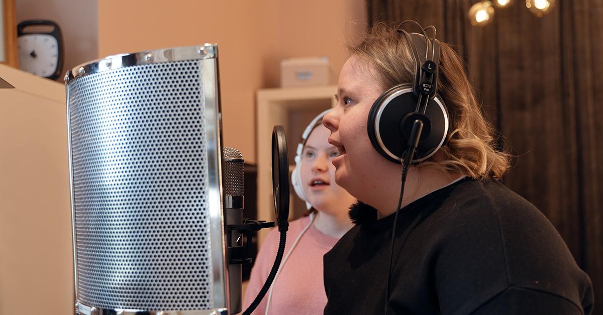 Två kvinnor med hörlurar sjunger i stor mikrofon.