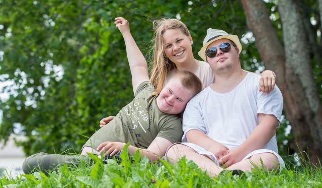 En ung kivnna och två unga killar med Downs syndrom sitter i gräset och håller om varandra.